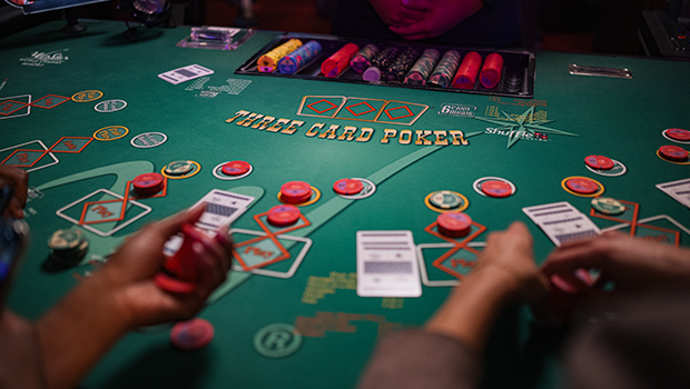 A close-up of a dealer’s hands distributing cards on a green Three Card Poker table with stacks of betting chips.