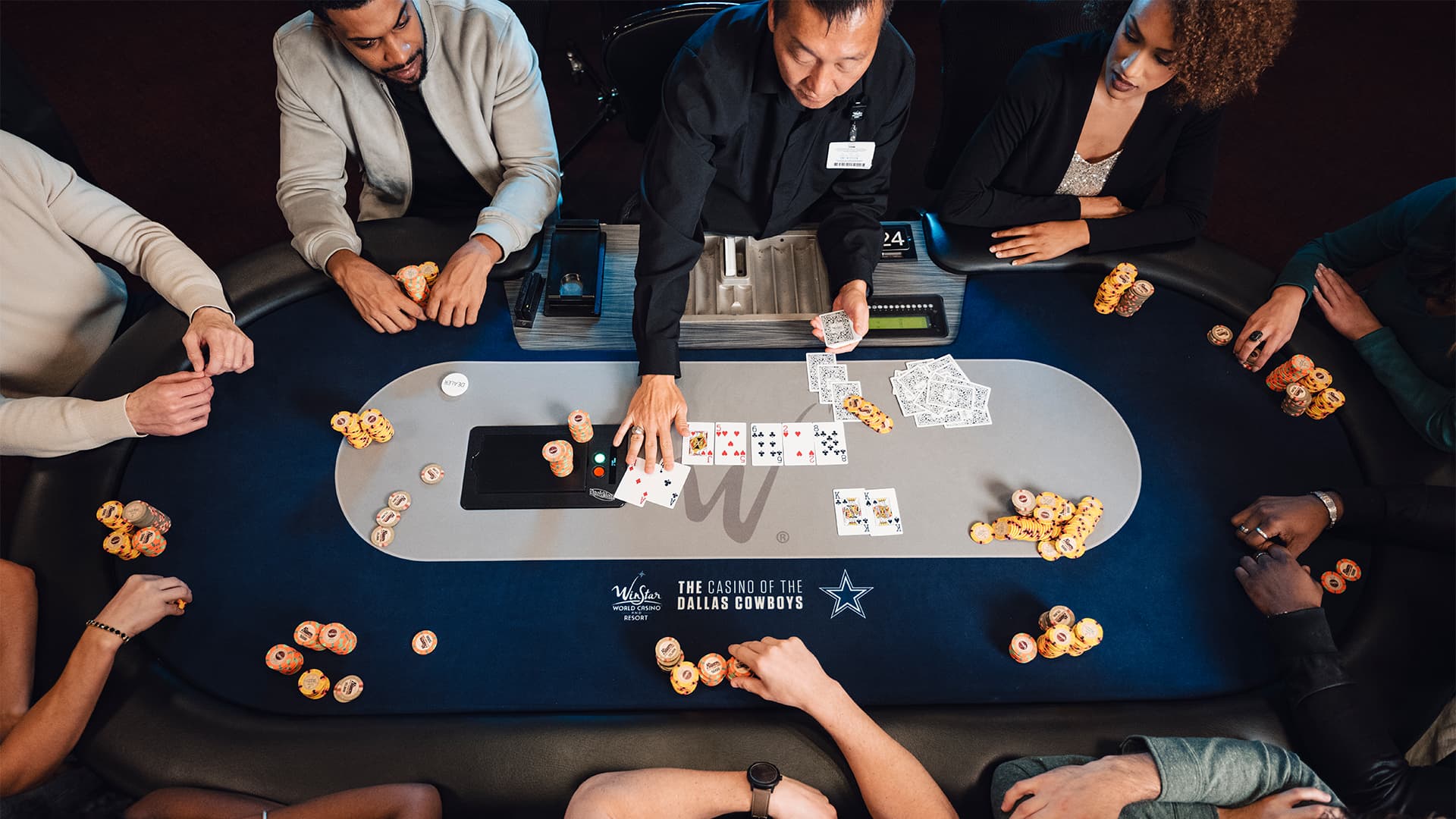 A group of people dressed in semi-formal attire playing a high-stakes poker game at a professional blue felt table inside a casino poker room.