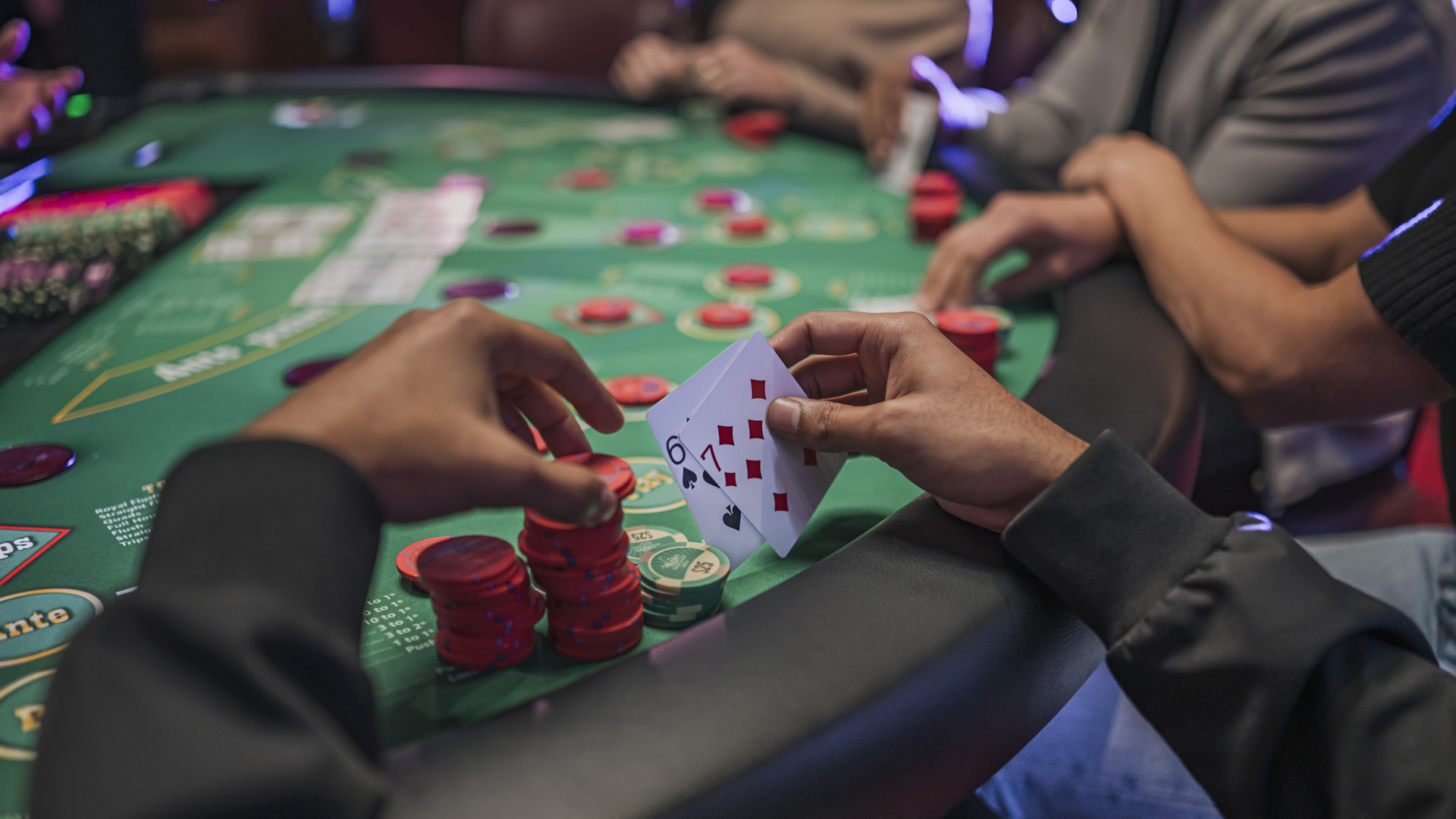 A player's hands holding red poker chips and two cards face-down on a green felt Ultimate Texas Hold'em table